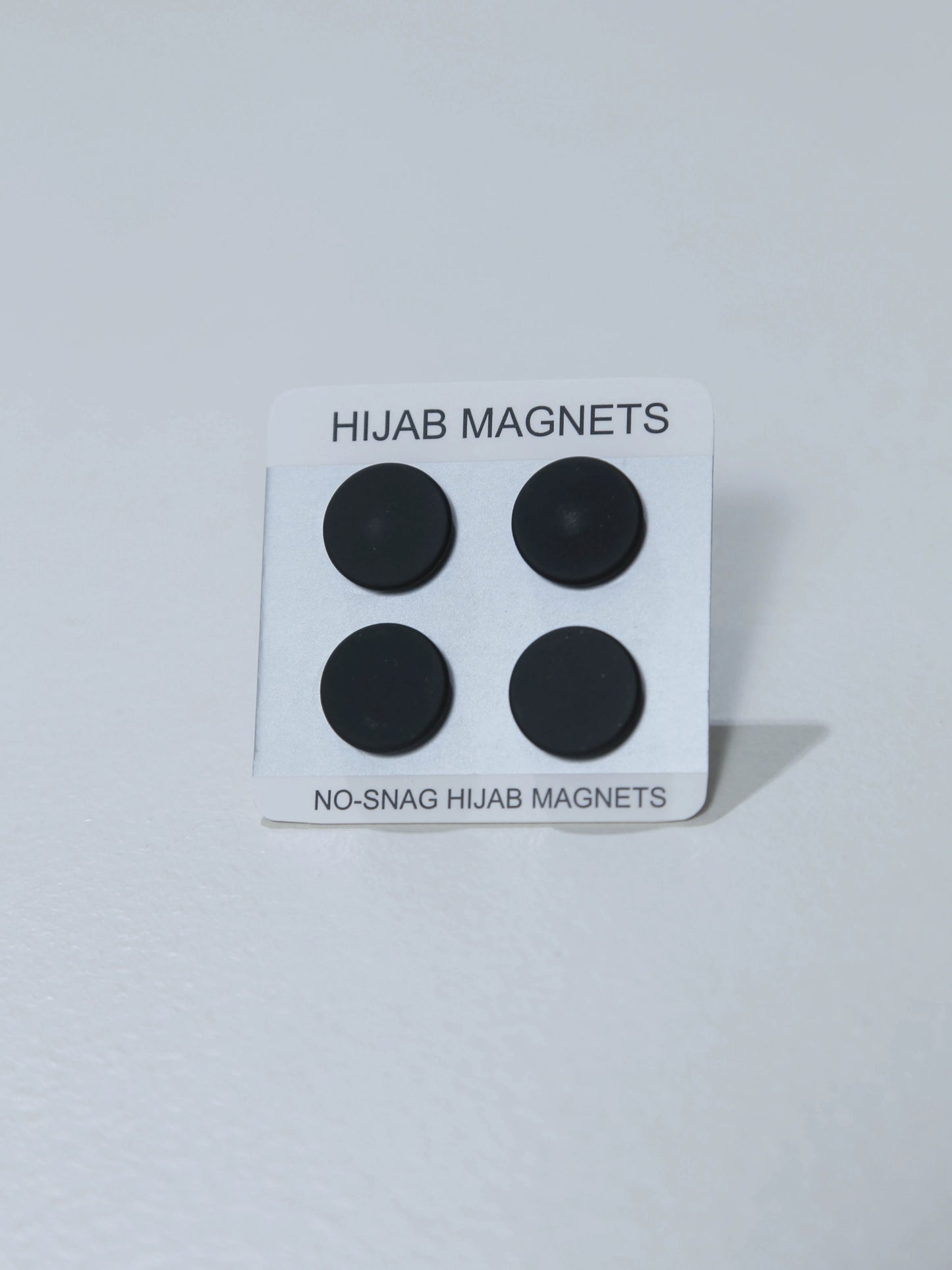 Packaging of hijab magnets with four black circular magnets on a white background.
