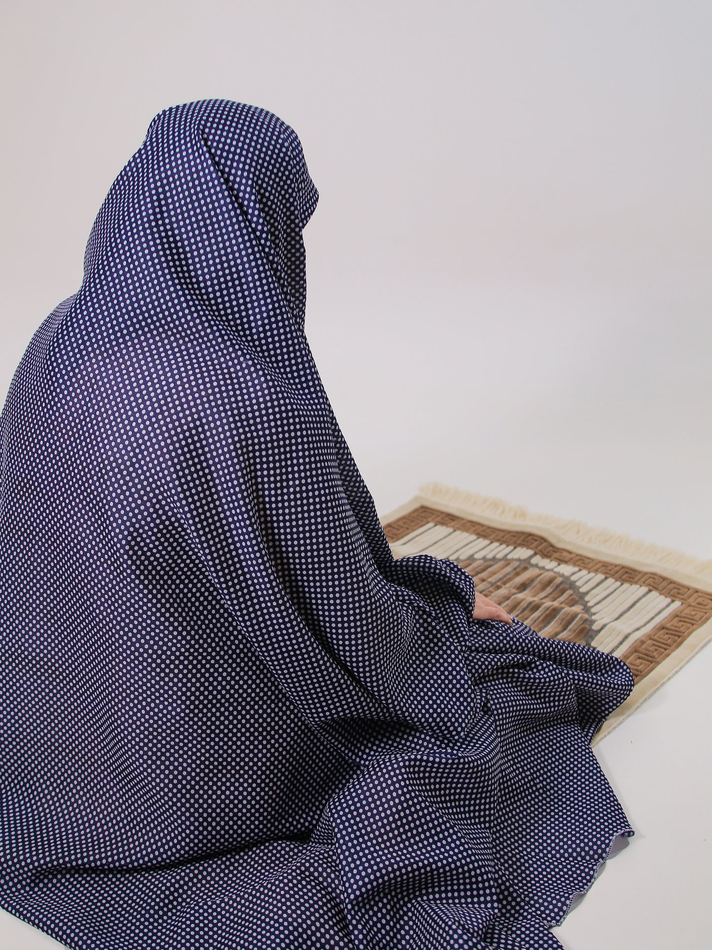 Person wearing a blue checkered abaya sitting on a prayer mat against a white wall.