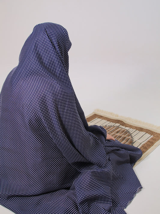 Person wearing a blue checkered abaya sitting on a prayer mat against a white wall.