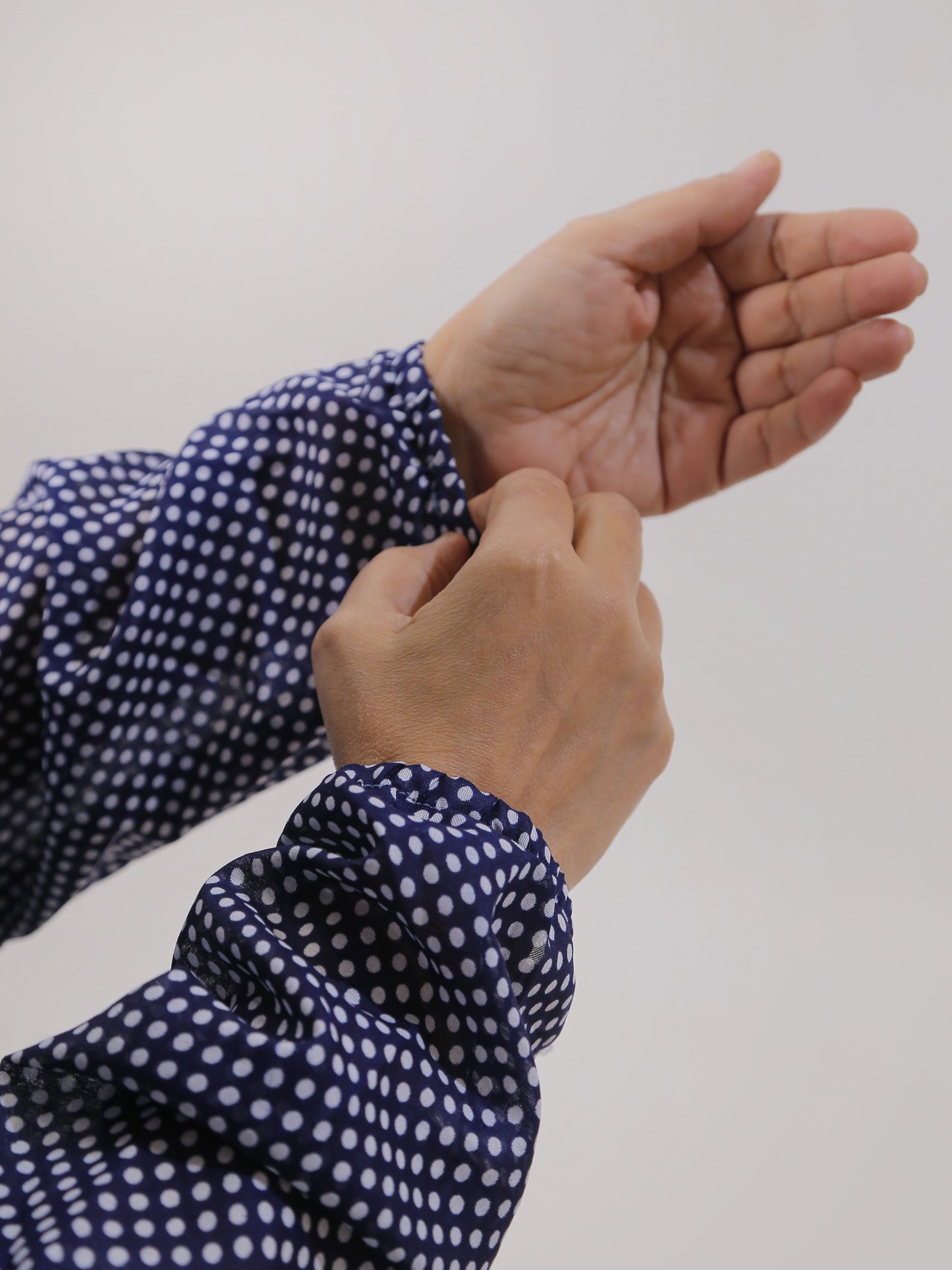 Person wearing a navy blue shirt with white polka dots, adjusting the sleeve on a plain background.