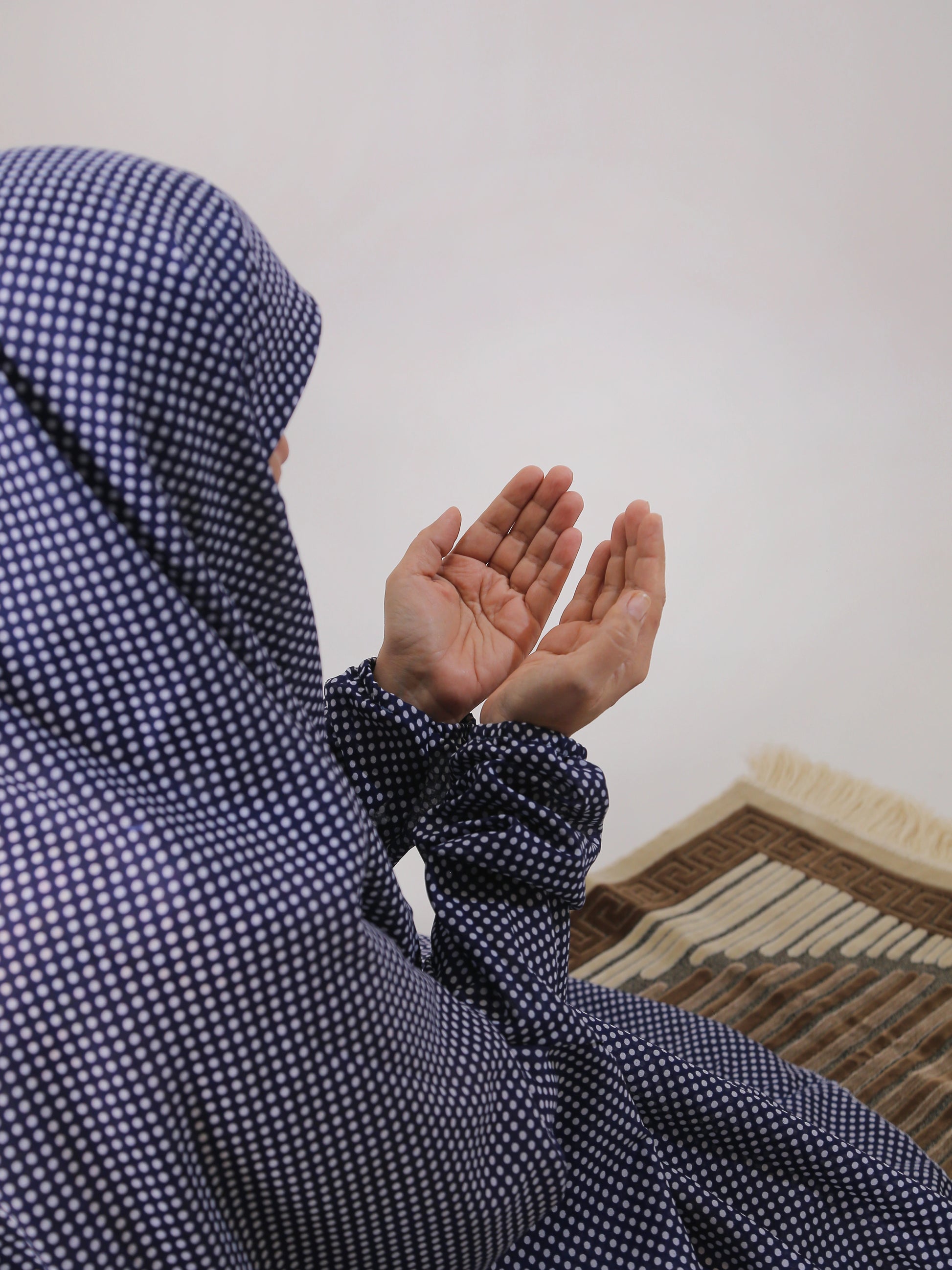 Person wearing a blue checkered hijab sitting on a mat with hands raised in a gesture of prayer or supplication.