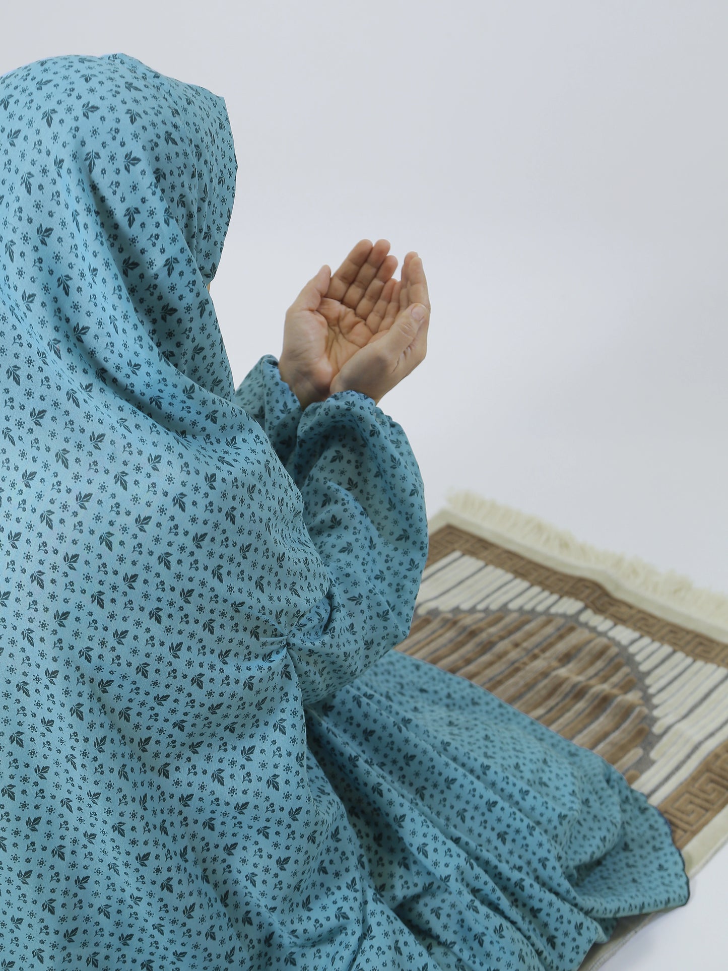 Person wearing a blue headscarf sitting on a patterned mat with hands raised.
