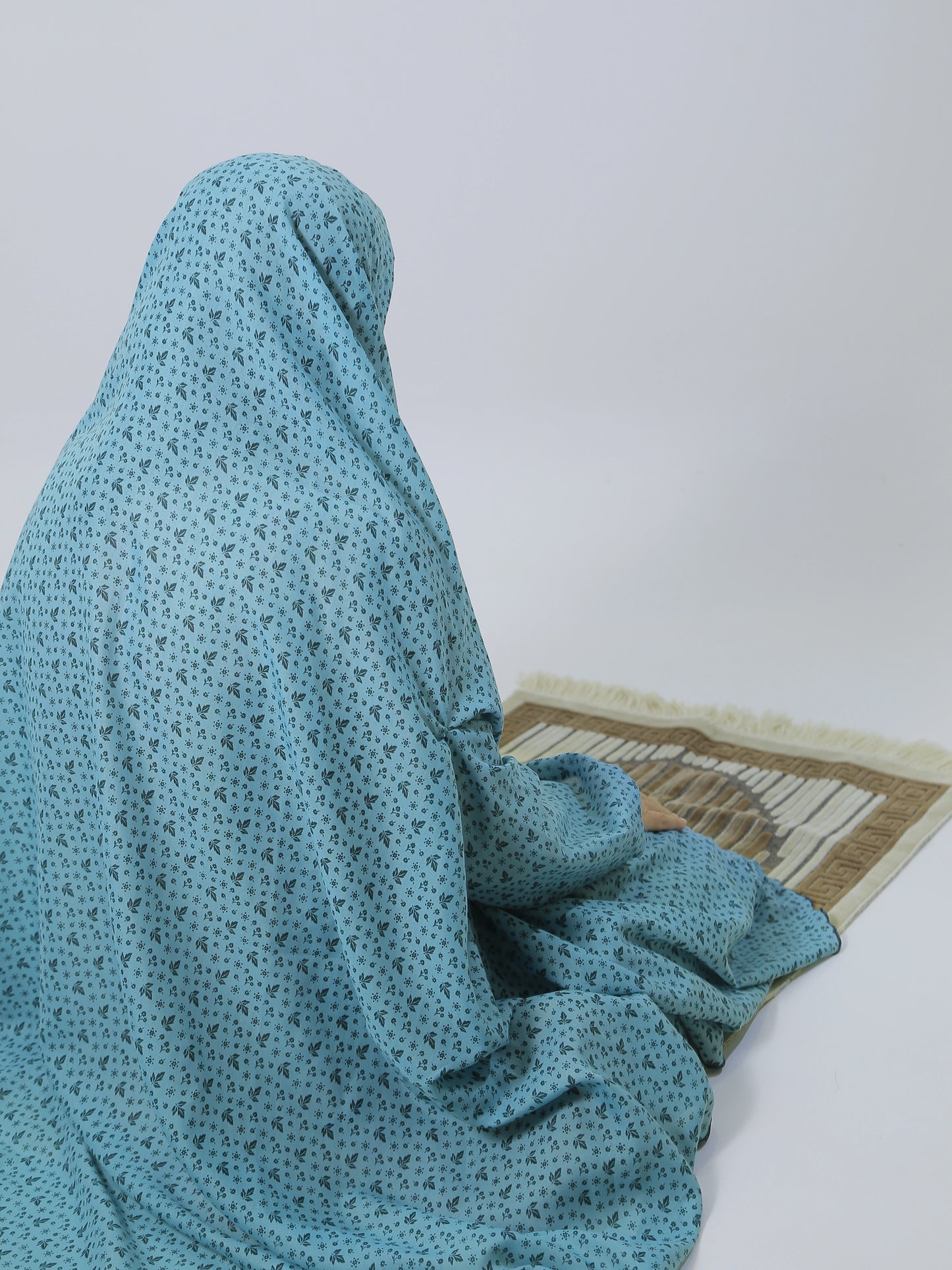 Person wearing a blue hijab sitting on a prayer mat against a white wall