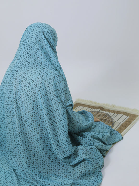 Person wearing a blue hijab sitting on a prayer mat against a white wall